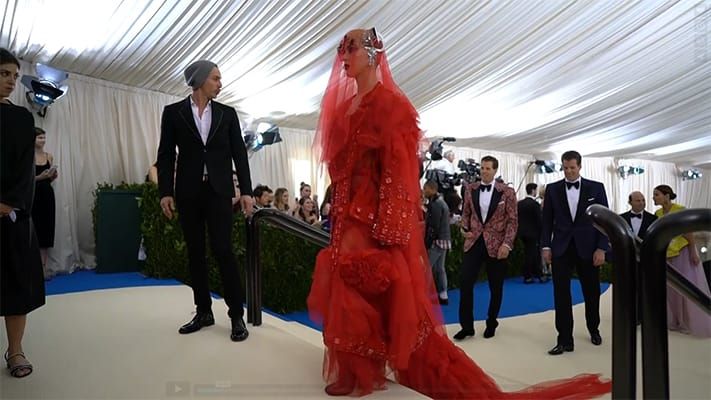 A woman in a haute couture, red dress with veil walking through a tented enclosure with people in evening wear following