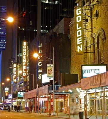 Nighttime photo of a New York City Street with neon signs displaying the names of theaters
