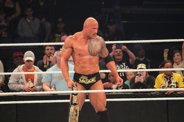 A well-muscled man in a wrestling ring wearing wrestling shorts & holding a championship belt glowering at someone off-camera