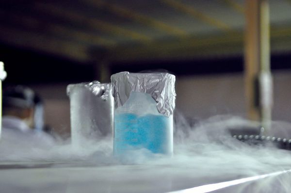 A photo of a pale blue liquid in a beaker, surrounded by gray vapor, a second (empty) beaker is in the background
