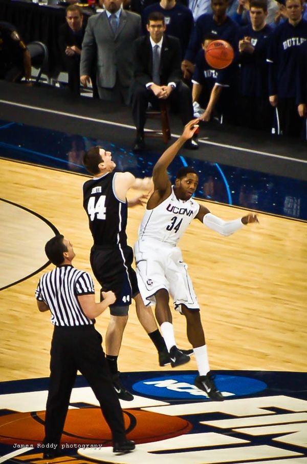 Two basketball players leap to get the opening tip of a match; a referee looks on