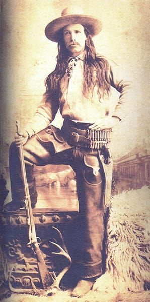 B&W photo of a long-haired, mustachioed man in a wide-brimmed hat, with a six-gun holstered at his side and holding a rifle
