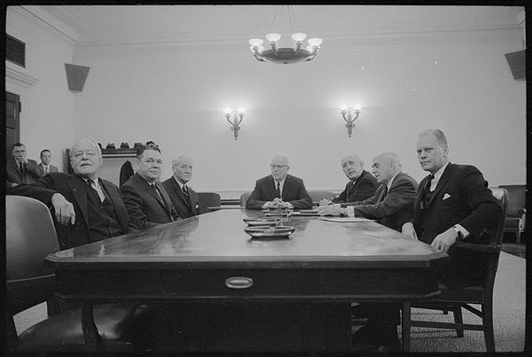 B&W photo of seven men in suits sitting around a conference table.
