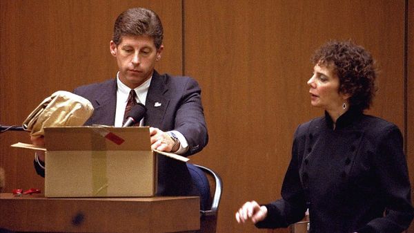 Photo of a man in a suit in a witness stand examining the contents of a box; a woman next to him is speaking to him