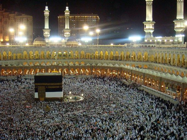 Nighttime photo of a large black cube surrounded by pilgrims within an open-air mosque; beyond are minarets & city buildings