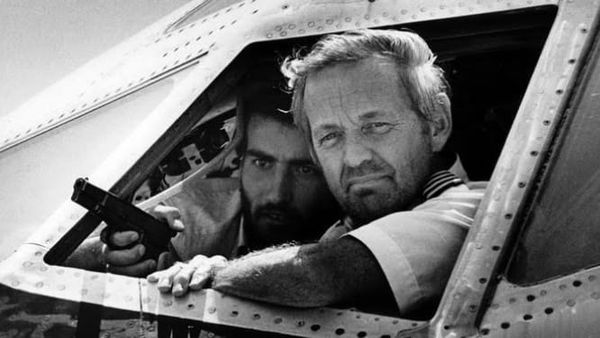 B&W photo of two men looking out the window of a airliner cockpit, one in a pilot’s uniform, the other holding a pistol