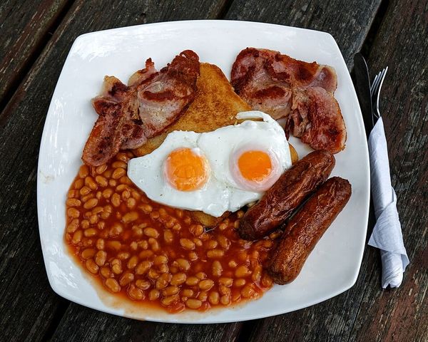 A breakfast plate containing two eggs, sausages, bacon, baked beans, and fried bread