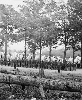B&W photo of a company of soldiers standing at attention in front of a line of trees with tents in the background