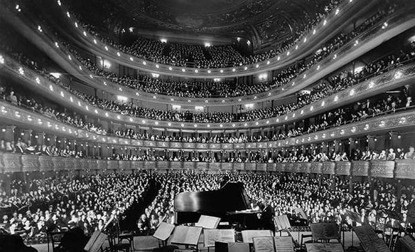 B&W photo taken from the back of the stage of an audience seated in a grand opera house; the stage is set for a piano concert