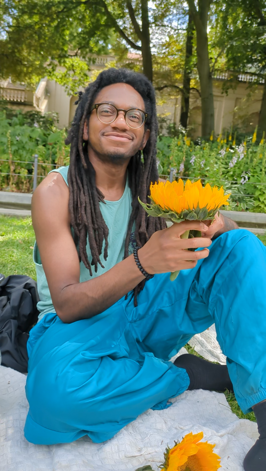 Dera is sitting on a white blanket in a park, smiling softly as he looks past the camera. He holds  a large sunflower, and there's another on the blanket. He is dressed in shades of green and blue, and his locs reach his biceps