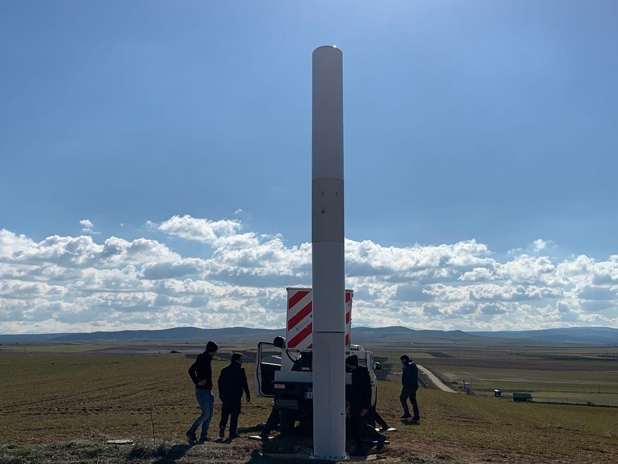 Tapered white cylindrical object 8 metres tall, in front of workers and truck, background of treeless plain and distant hills