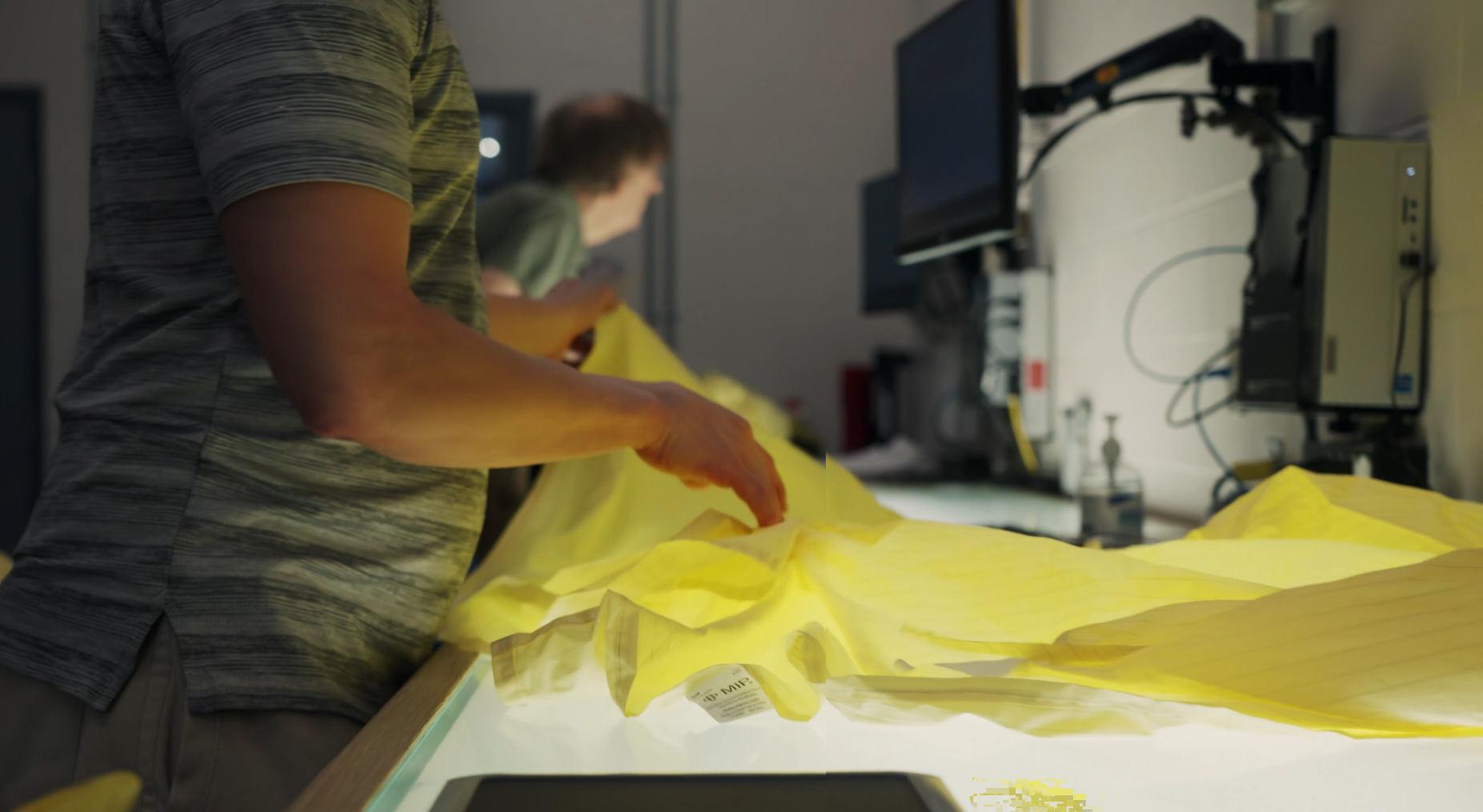 Photo of a worker passing a yellow hospital isolation gown over a light table to inspect it for defects.