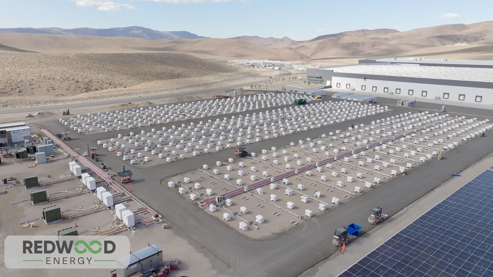 Aerial photo of parking lot-sized array of white battery modules adjacent to a photovoltaic array and large building