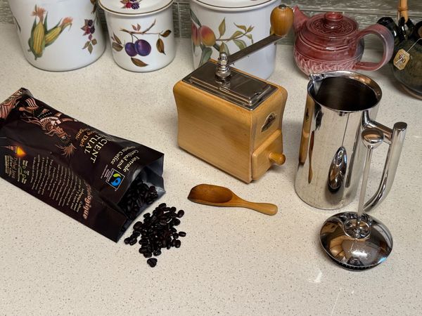Kitchen counter with open bag of coffee beans, on its side with beans spilling out, next to manual coffee grinder and French press. Behind are ceramic canisters and teapots.