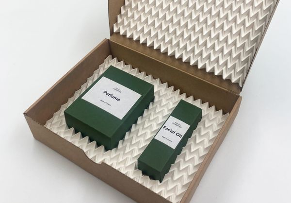 Open cardboard box with attached lid, containing two smaller green cosmetics boxes cradled in protective packaging made of machine-made, custom-fitted paper origami