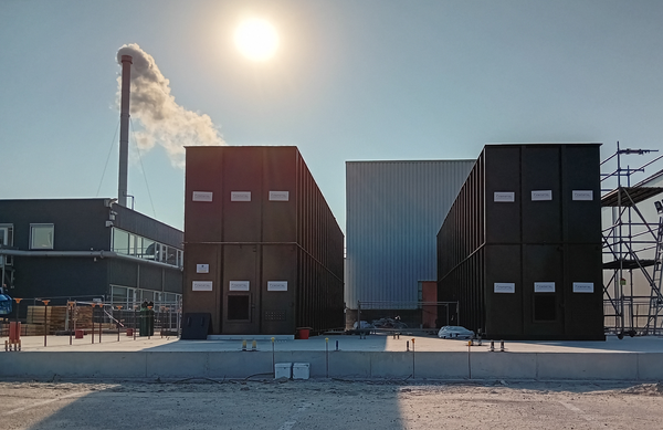 A pair of two-story tall rectangular brown high temperature heat storage containers next to a boiler house, whose chimney contains a boiler that the storage will replace