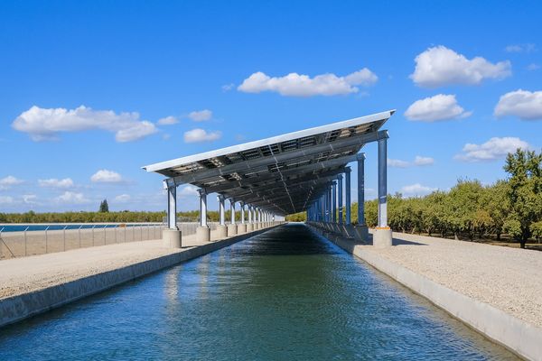 Sunnay day photo of narrow irrigation canal covered with solar photovoltaic panels, alongside orchards