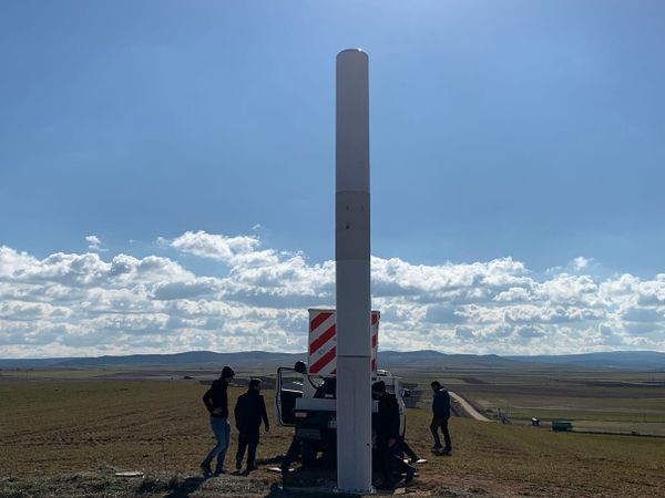 Tapered white cylindrical object 8 metres tall, in front of workers and truck, background of treeless plain and distant hills