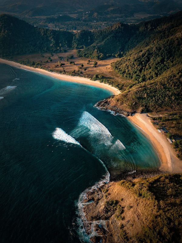 Aerial view of a temperate coastline showing clear blue ocean water and clean white breakers rolling into a little bay.