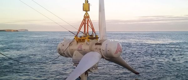 Three-bladed tidal turbine being lowered into place by a large crane into the sea off Scotland's Pentland Firth, cliffs in the background.