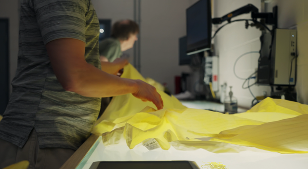Photo of a worker passing a yellow hospital isolation gown over a light table to inspect it for defects.