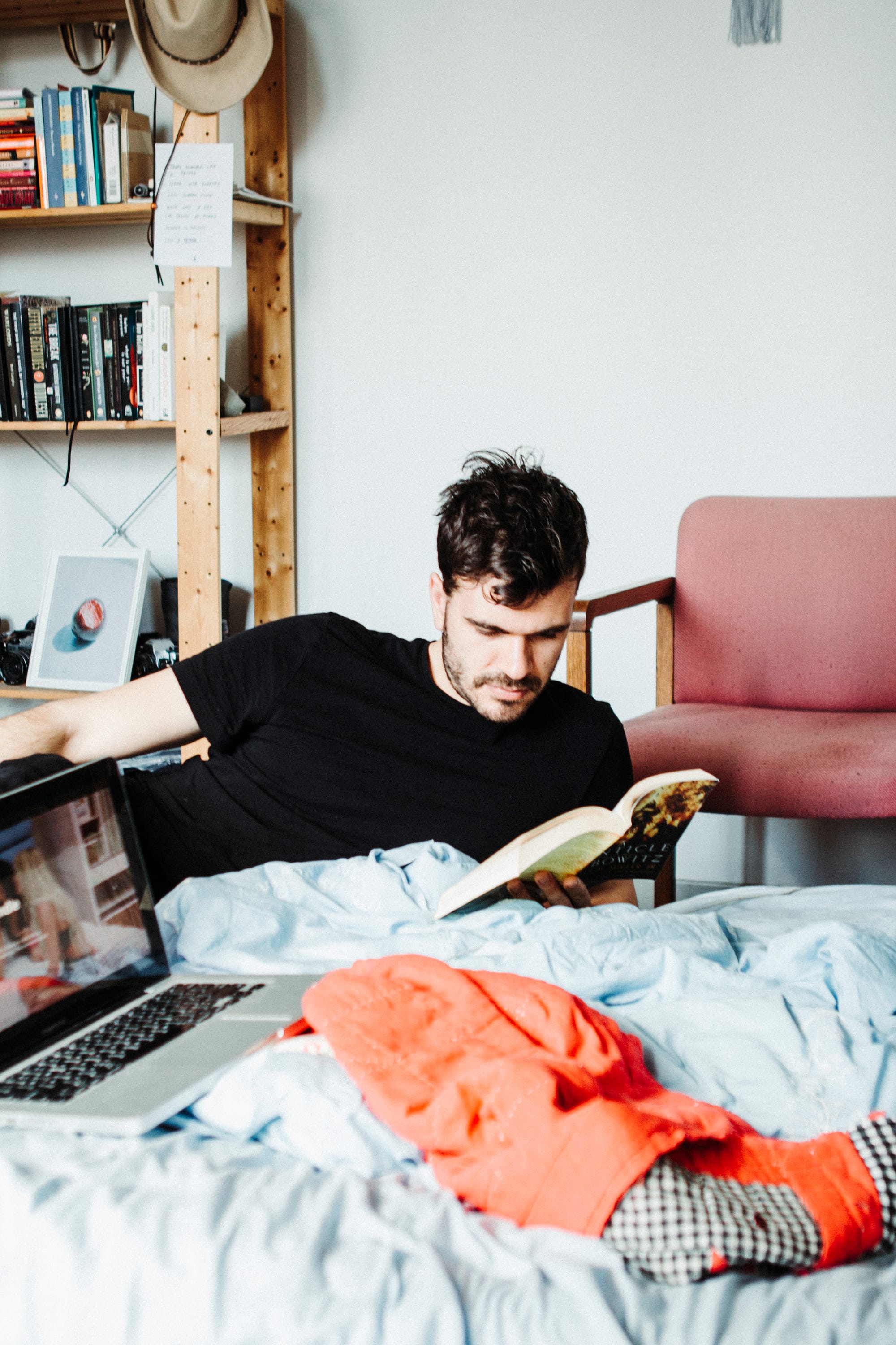 Photograph shows Alyson's ex lying on her bed reading. A pink chair and bookshelf is in the background. In the foreground is a red blanket and a open laptop. 