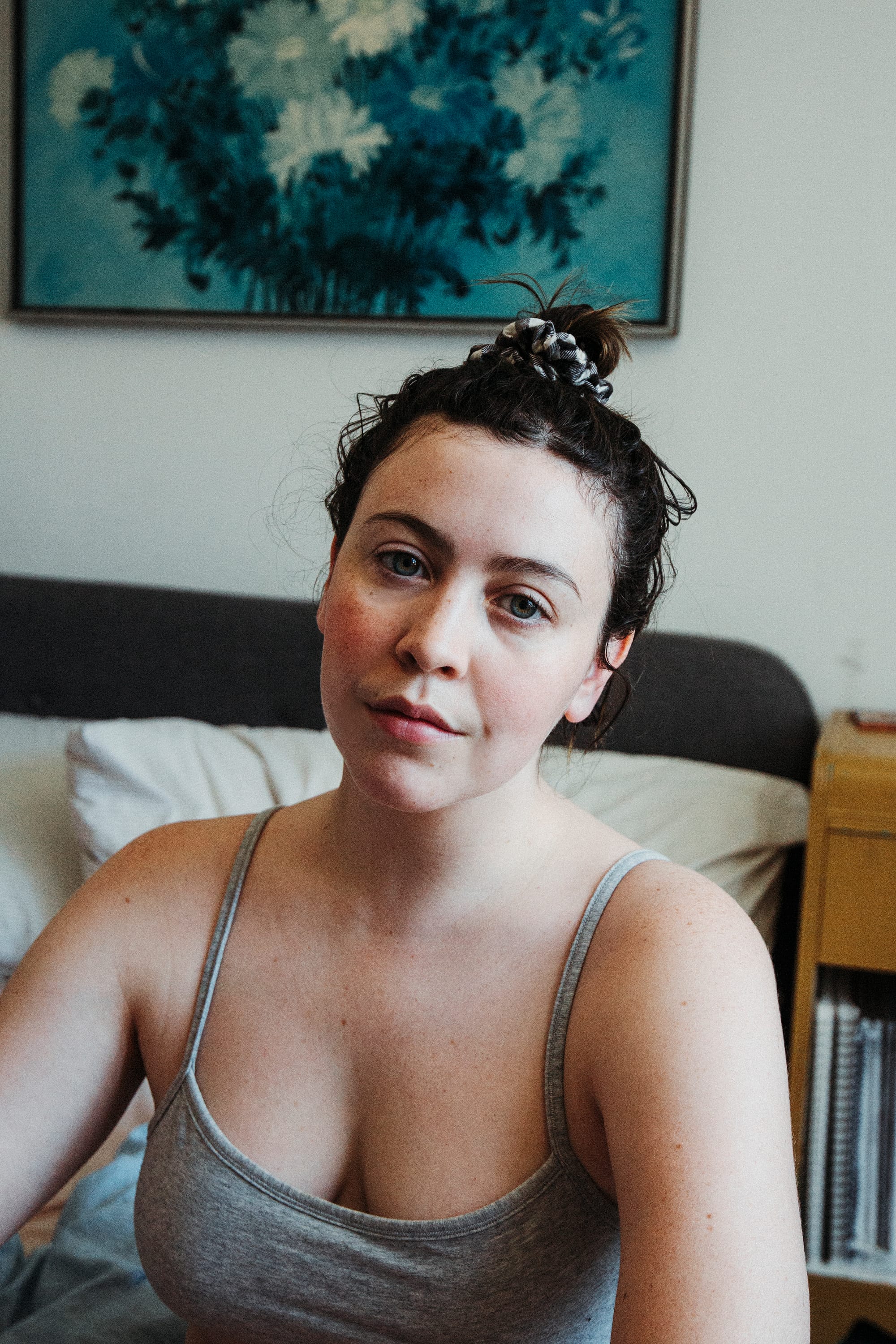 A self portrait of Alyson: a White woman with blue eyes is wearing a grey bra top and revealing her freckly skin. She has dark brown hair up in a bun and sits on her bed, which is in the background out of focus, along with her yellow side table. A blue floral painting hung on the wall is shown at the top of the frame behind Alyson..