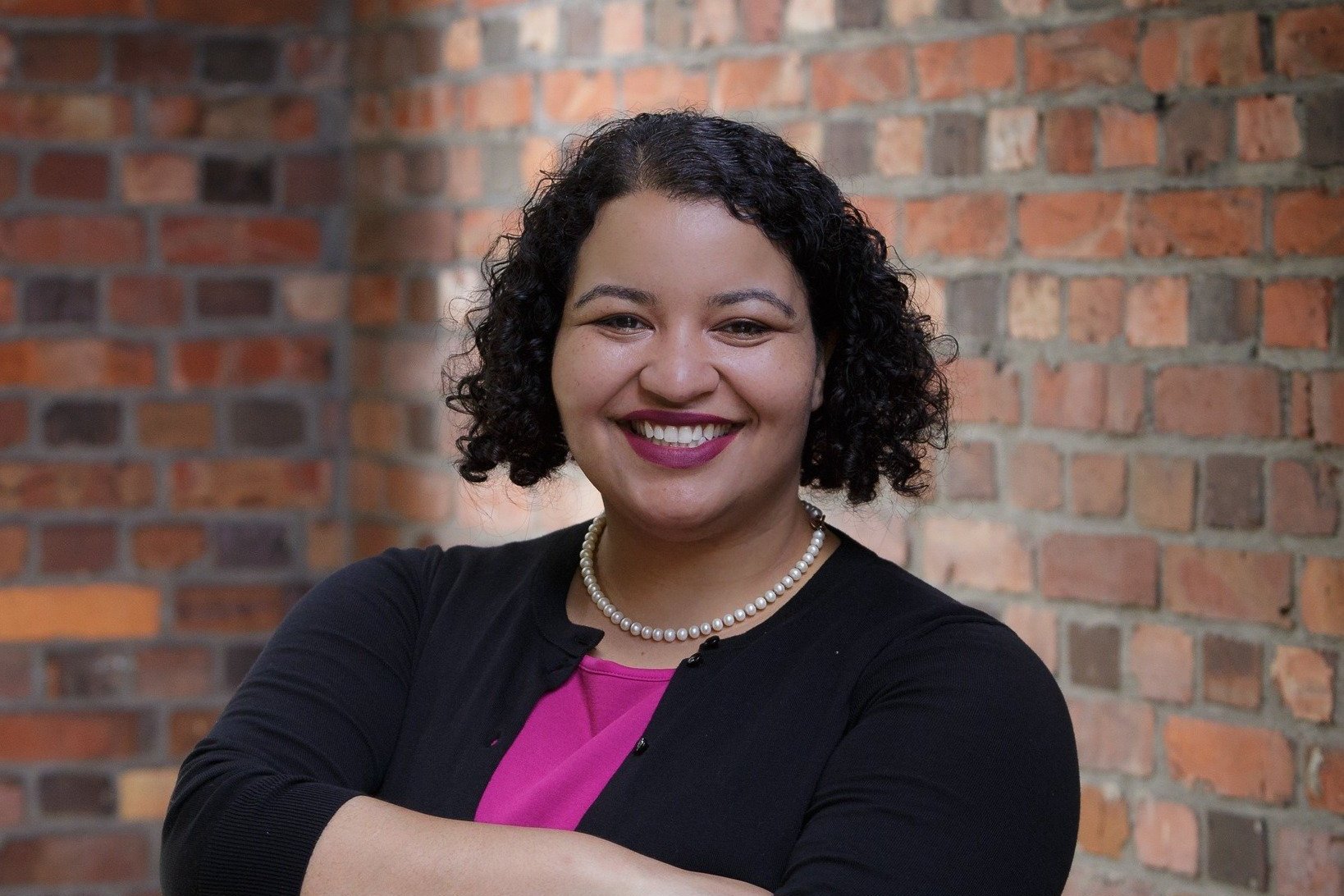 A portrait of Julia Reed, smiling towards the camera, standing infront of a brick wall.