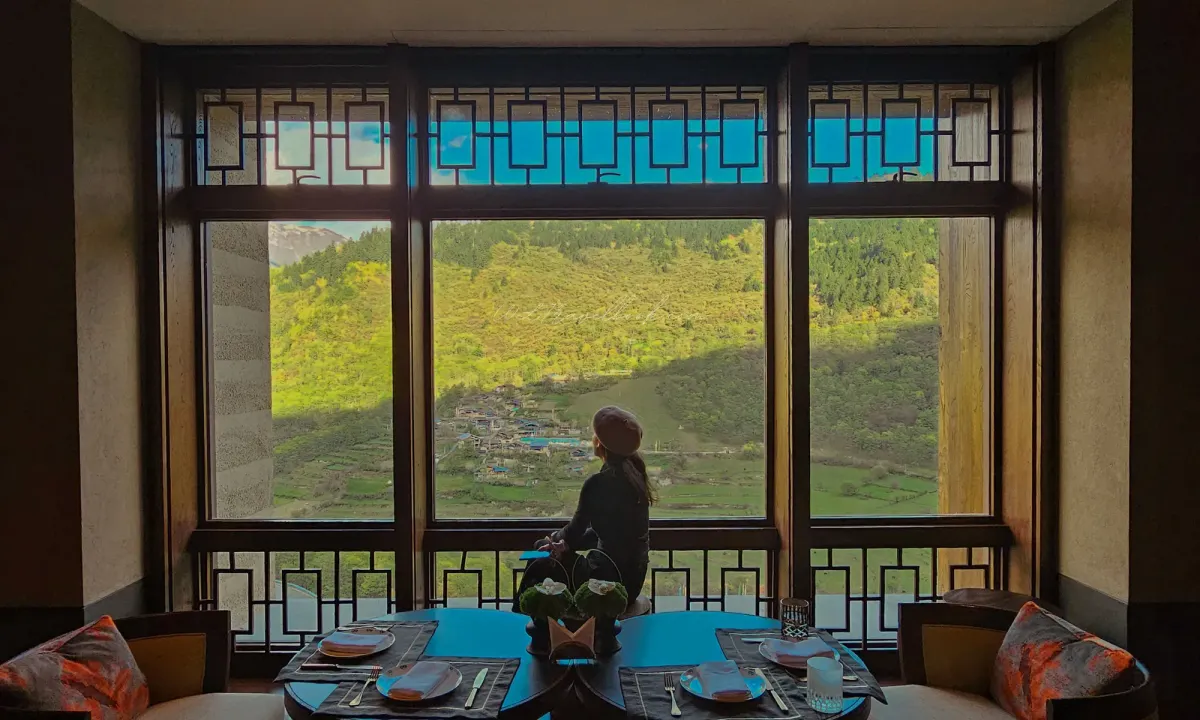 Figure in blue sitting at interior window of Rissai Valley overlooking golden snow mountain landscape through architectural framing, by WLTravelbook