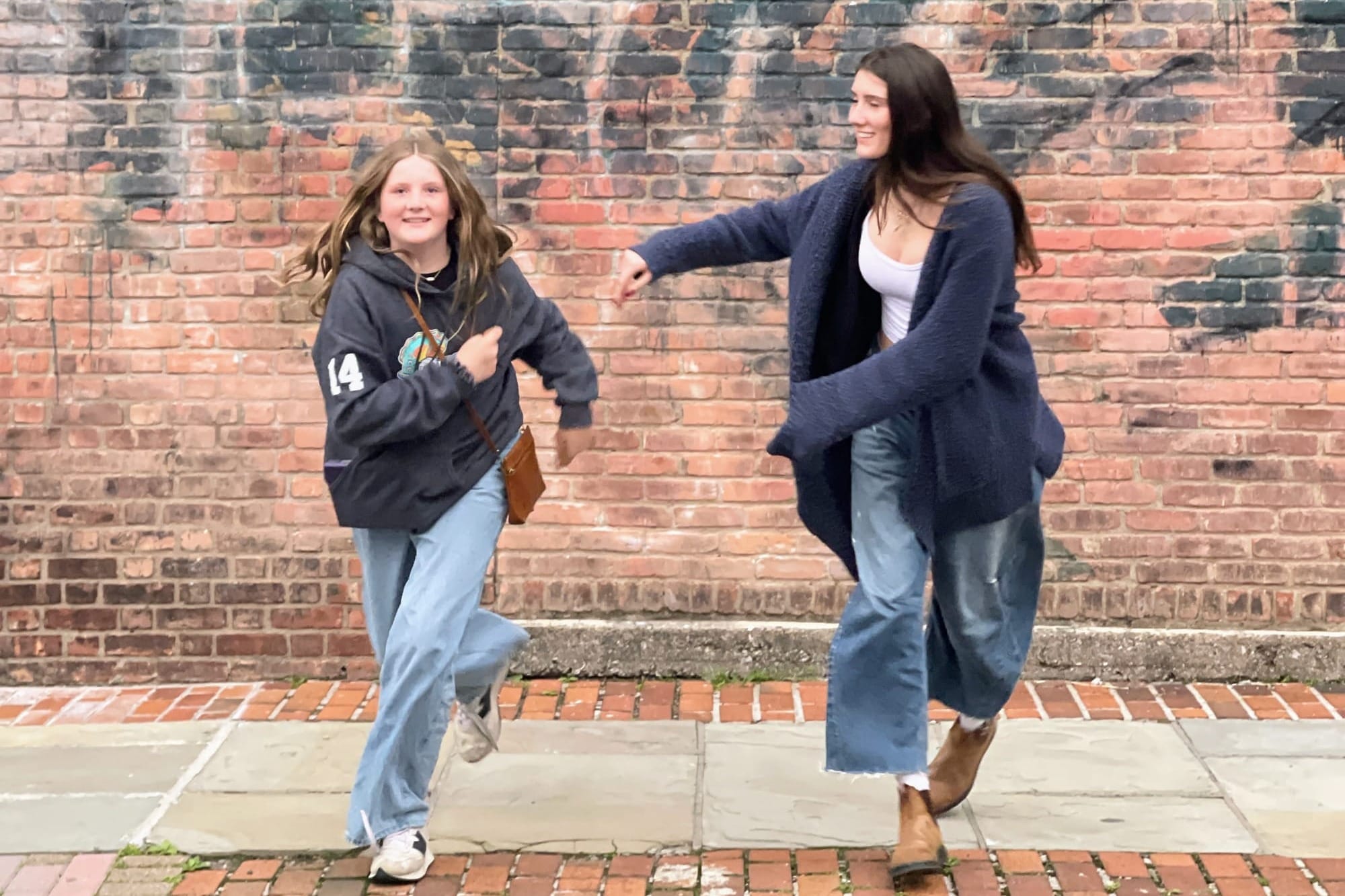 Two girls wearing jeans and blue tops run towards the camera, away from a graffiti red brick wall