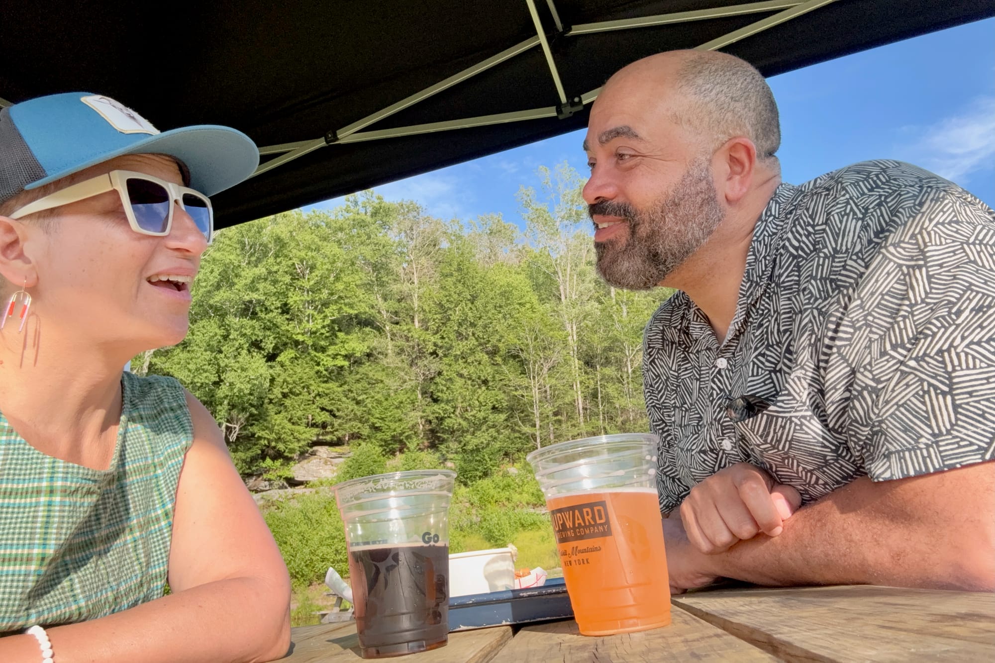 A woman in a blue ball cap and a man in a geometric shirt smile at each other over beers with trees in the background.