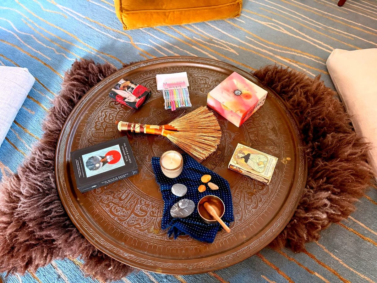 A brass Moroccan tray lies on top of a brown fur rug with oracle decks, a small broom, a candle, rocks and a singing bowl
