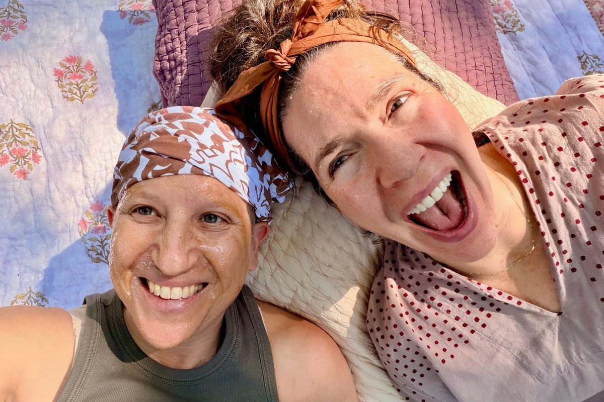 Two women in bandanas lie on a pink flower quilt with raw honey on their faces.