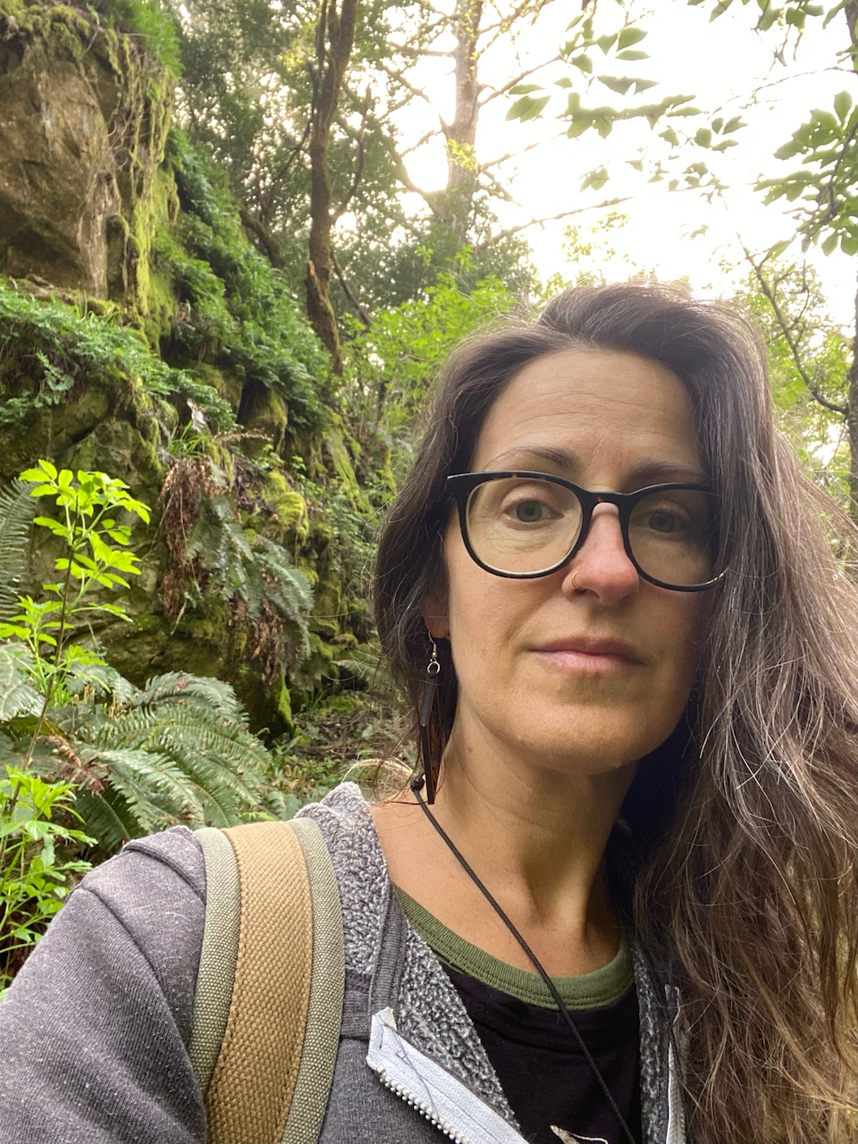 Woman with long brown and gray hair and glasses wears a backpack and grey hoodie on a lush hiking trail