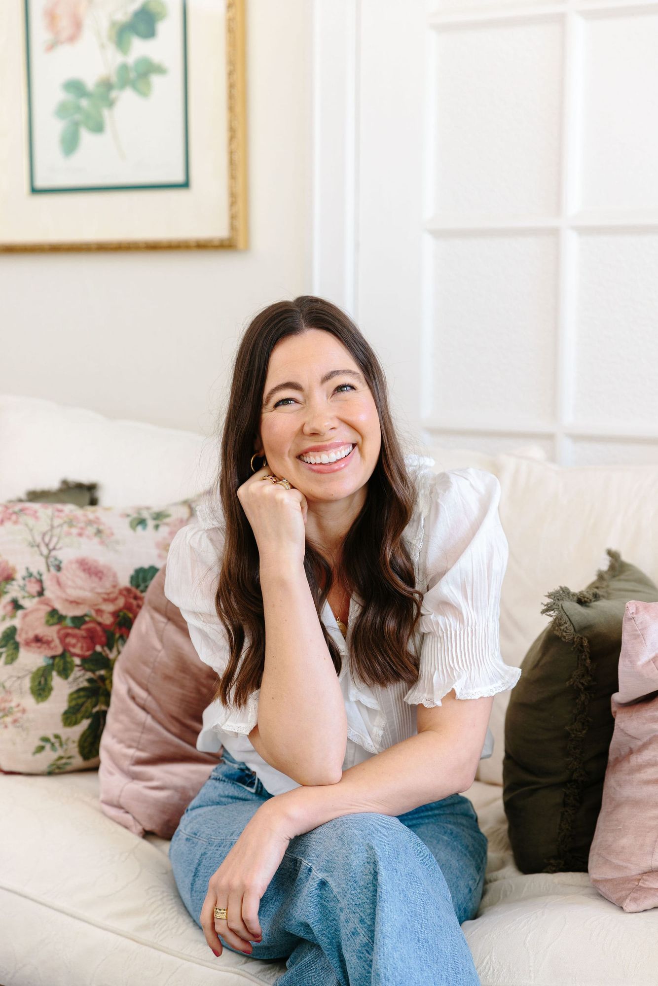 Woman with long brown hair smiling, wearing a white top and jeans, sitting on a leather couch with pink and green throw pillows