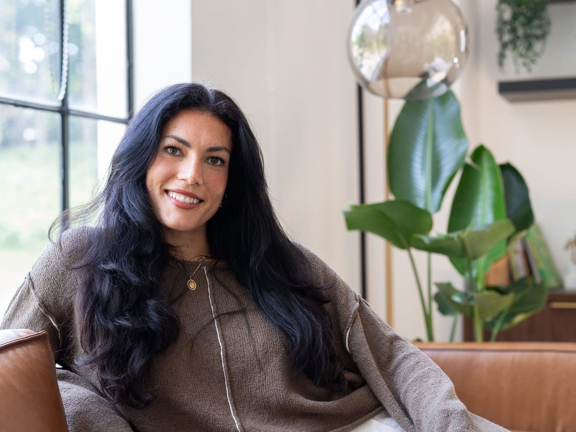 Woman with long dark hair and beige sweater sits on a tan leather couch in front of plants in a light-filled studio