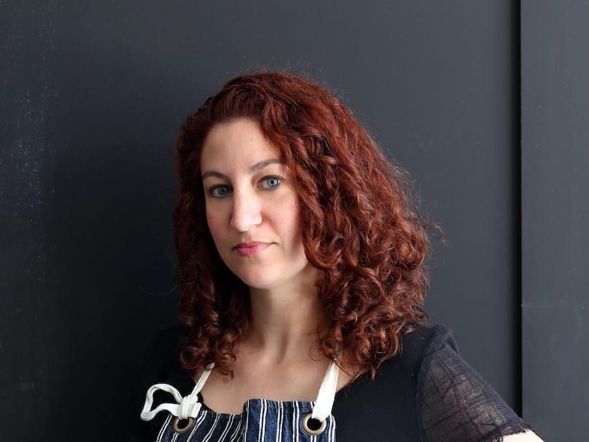 Woman with red curly hair in a dark shirt and apron stands against a dark grey door