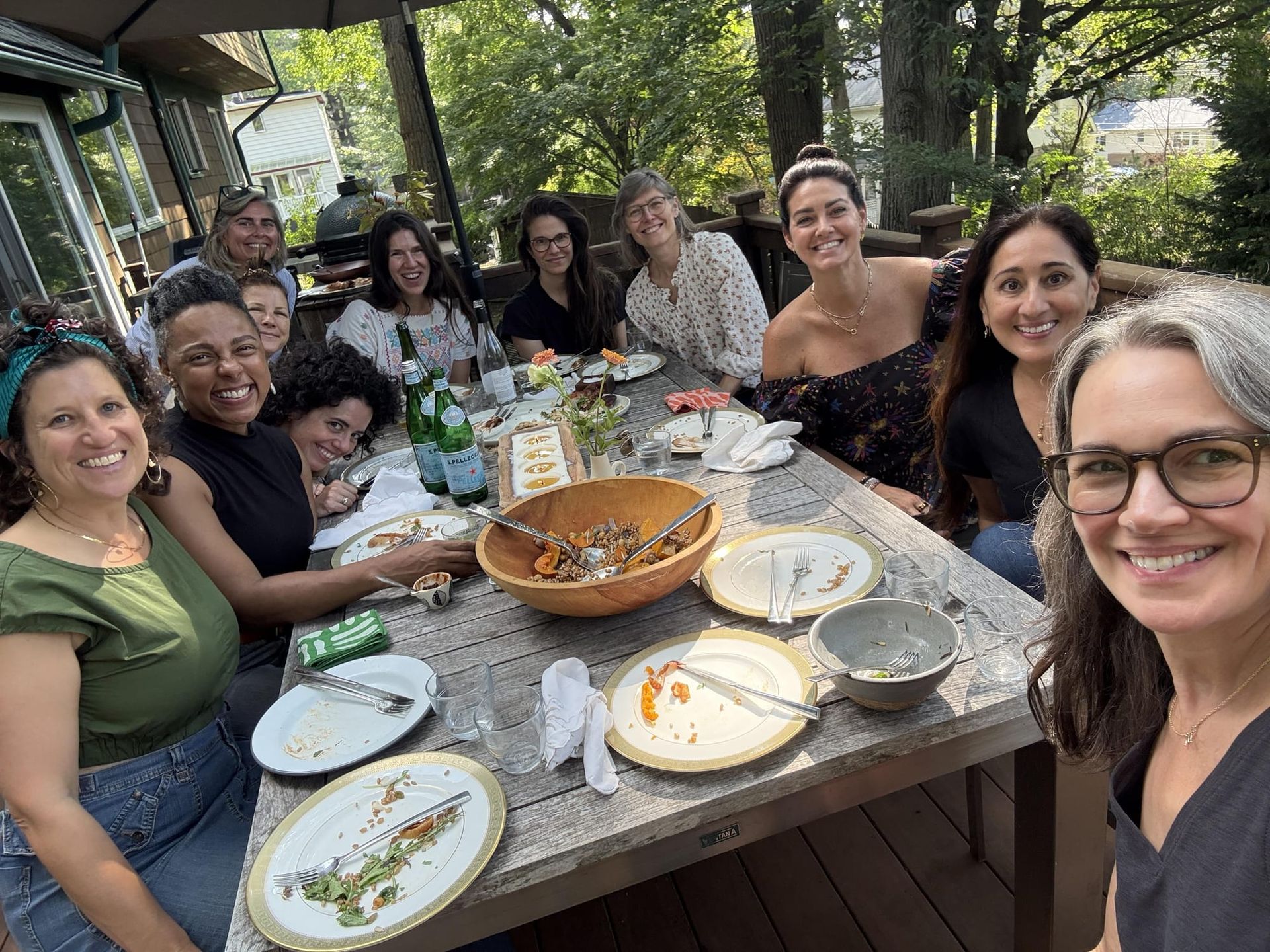 11 women gather around an outdoor picnic table with big smiles and only crumbs left on the plates after a shared meal