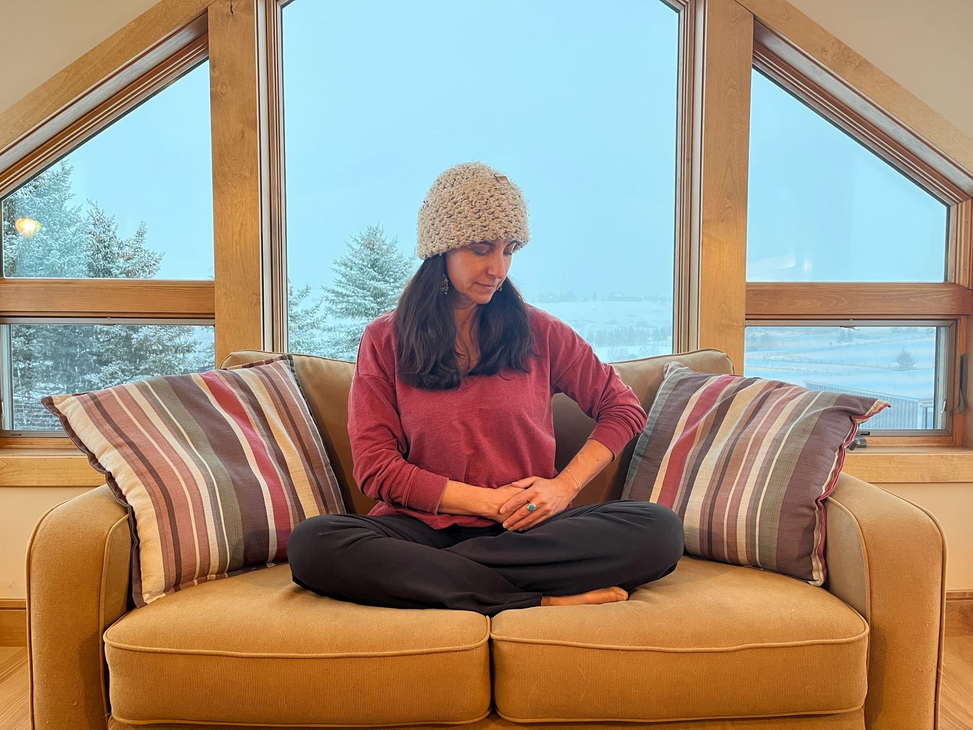 A woman sits atop her ochre-colored sofa, cross-legged in front of a large window with wintry trees in the distance