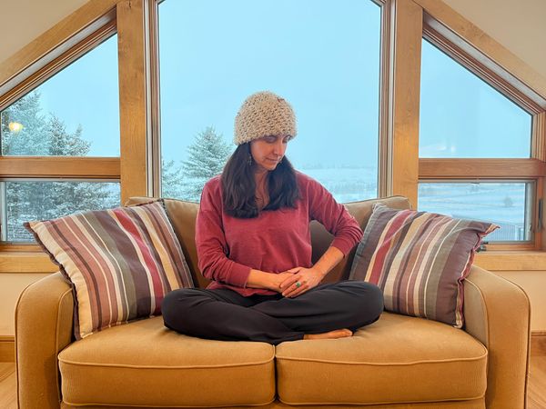 A woman sits atop her ochre-colored sofa, cross-legged in front of a large window with wintry trees in the distance