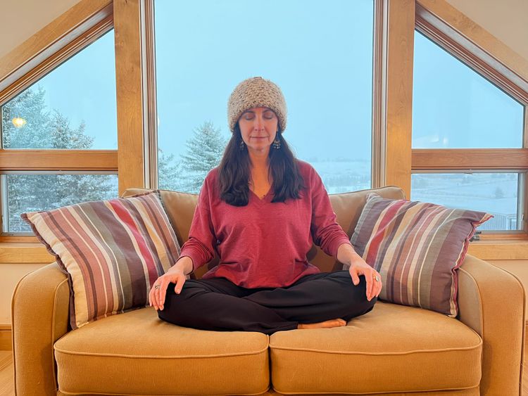 Woman meditating on a yellow couch in front of a window with snowy trees outside