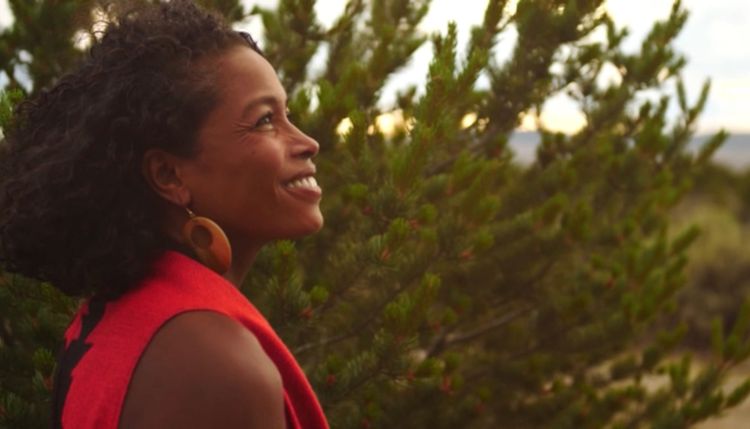 A black woman in a red top with large gold hoop earrings stands smiling in front of an evergreen bush