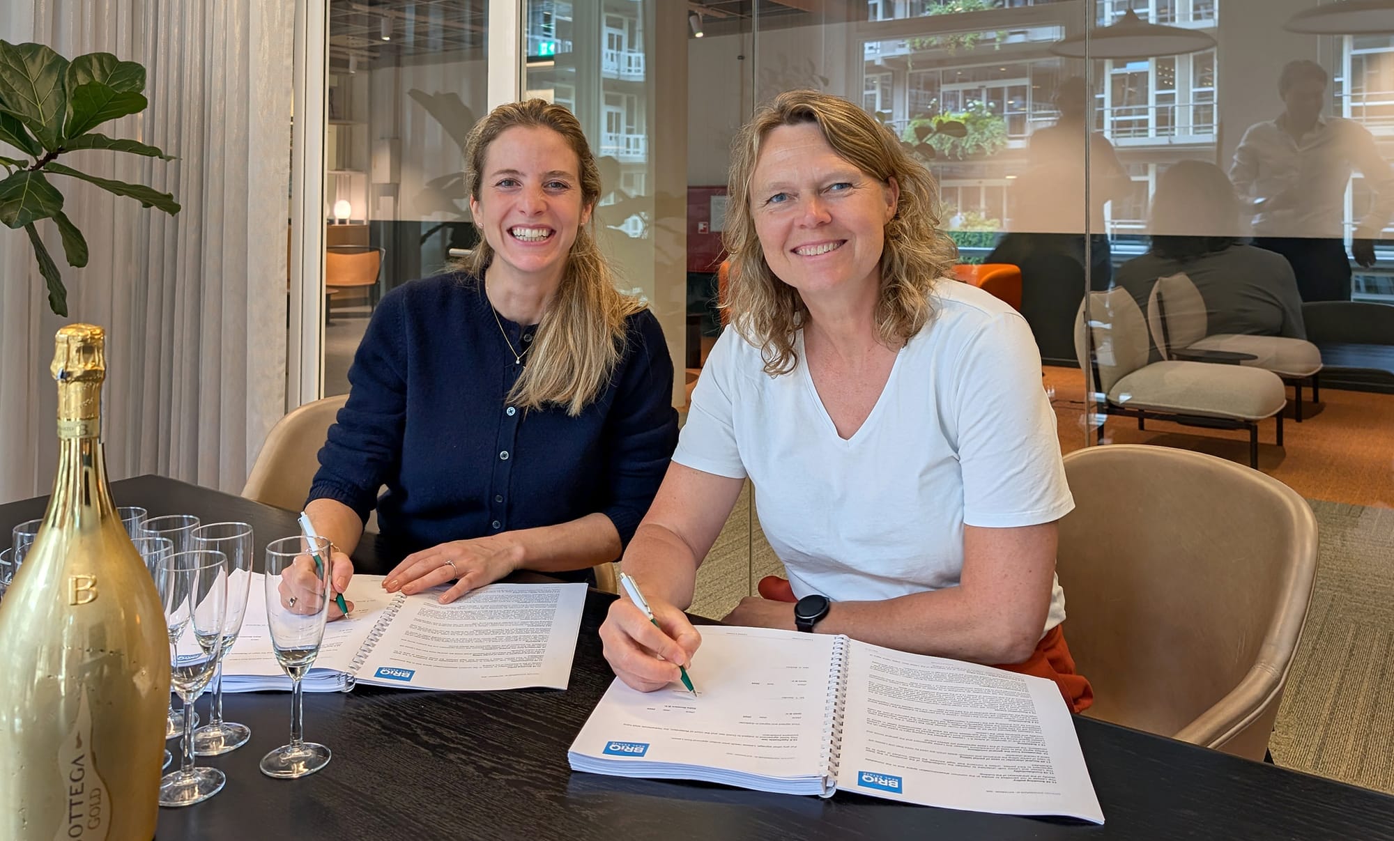 Twee glimlachende vrouwen die een document ondertekenen, zittend aan een tafel met champagneglazen.