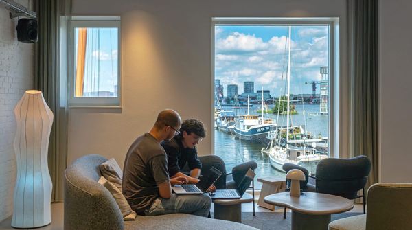Kantoorsetting met twee mannen op een bank met laptop op hun schoot, met op de achtergrond door een raam uitzicht op water en boten.