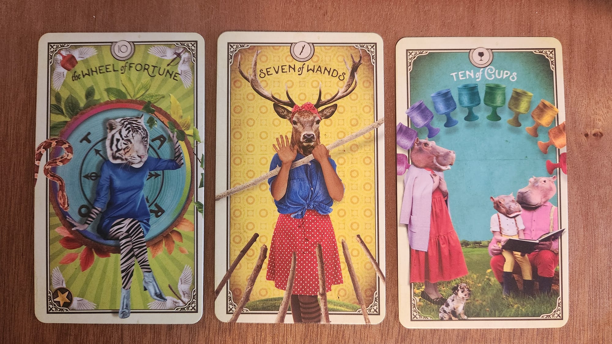 Wheel of fortune, drawn as a lady tiger sitting inisde a circle, a snake is just off to the side. Deer Lady holding a wand against 6 others in the middle, and two hippo parents with the father hippo reading a story to a child hippo beneath 10 cups in rainbow colours.