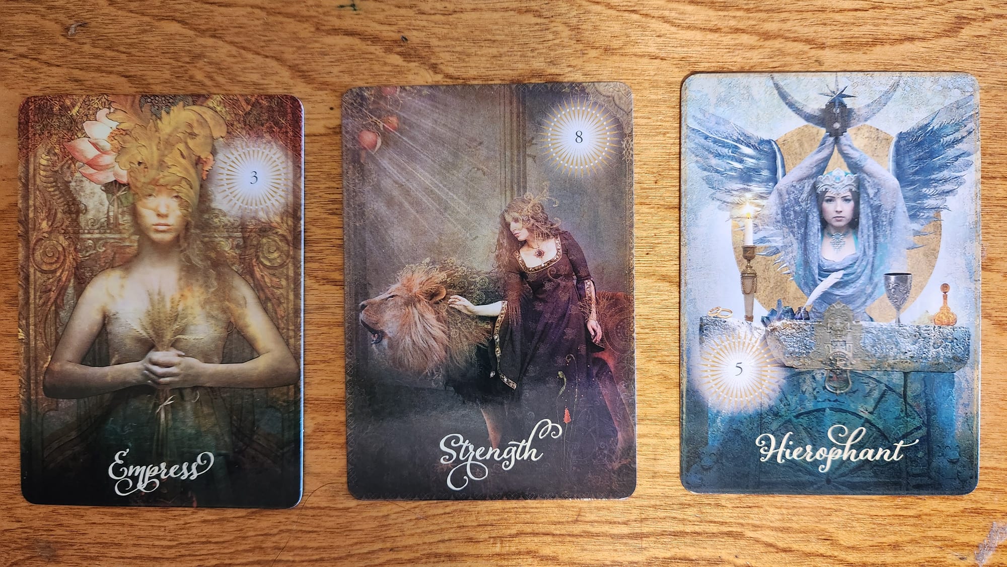 Three tarot cards. The empress holds a sheaf of wheat and has a headress of leaves and flowers. A woman in long medieval garb stands beside a roaring lion, her hand on it's mane. The Heirophant stands in front of an alter and holds a crescent moon above her head and has wings at her back.