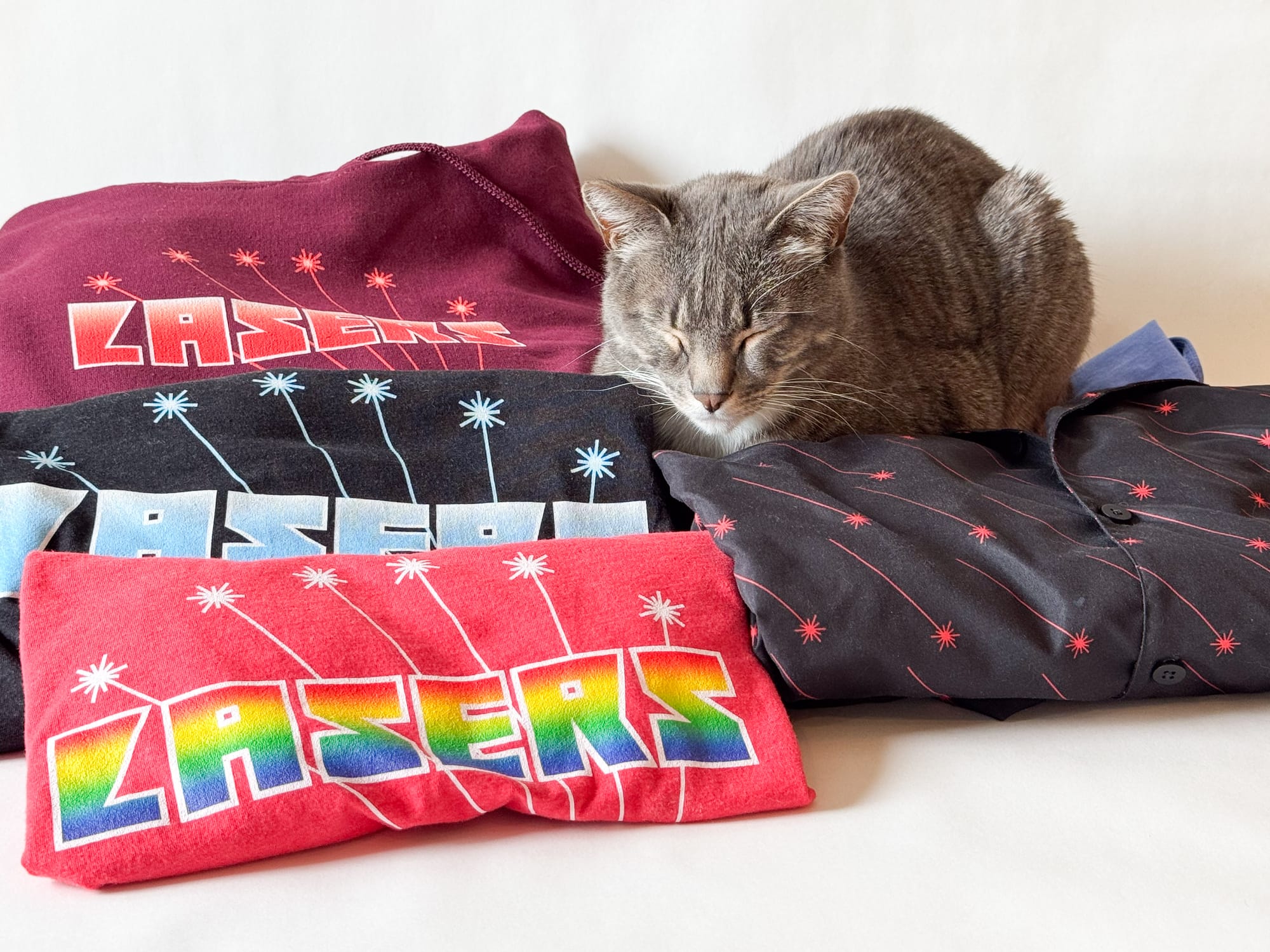 A grey tabby cat is in full cat-loaf mode, eyes closed and ears relaxed, while surrounded by three shirts that say "lasers" on them and one button-down shirt with a laser pattern.