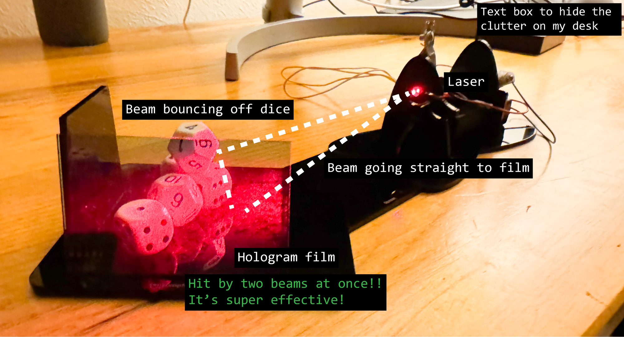 The setup is sitting on a plain wooden desk with a bunch of cords in the background. The laser is mostly visible as a red glow coming out of a nest of black plastic and wires. It illuminates a pile of six-sided and 12-sided dice and also makes a red glow on a glass plate of film that sits between the dice and the viewer.