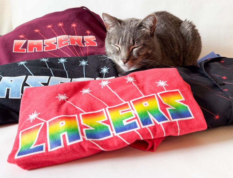 A grey tabby cat snoozes surrounded by folded shirts that say "lasers" on them