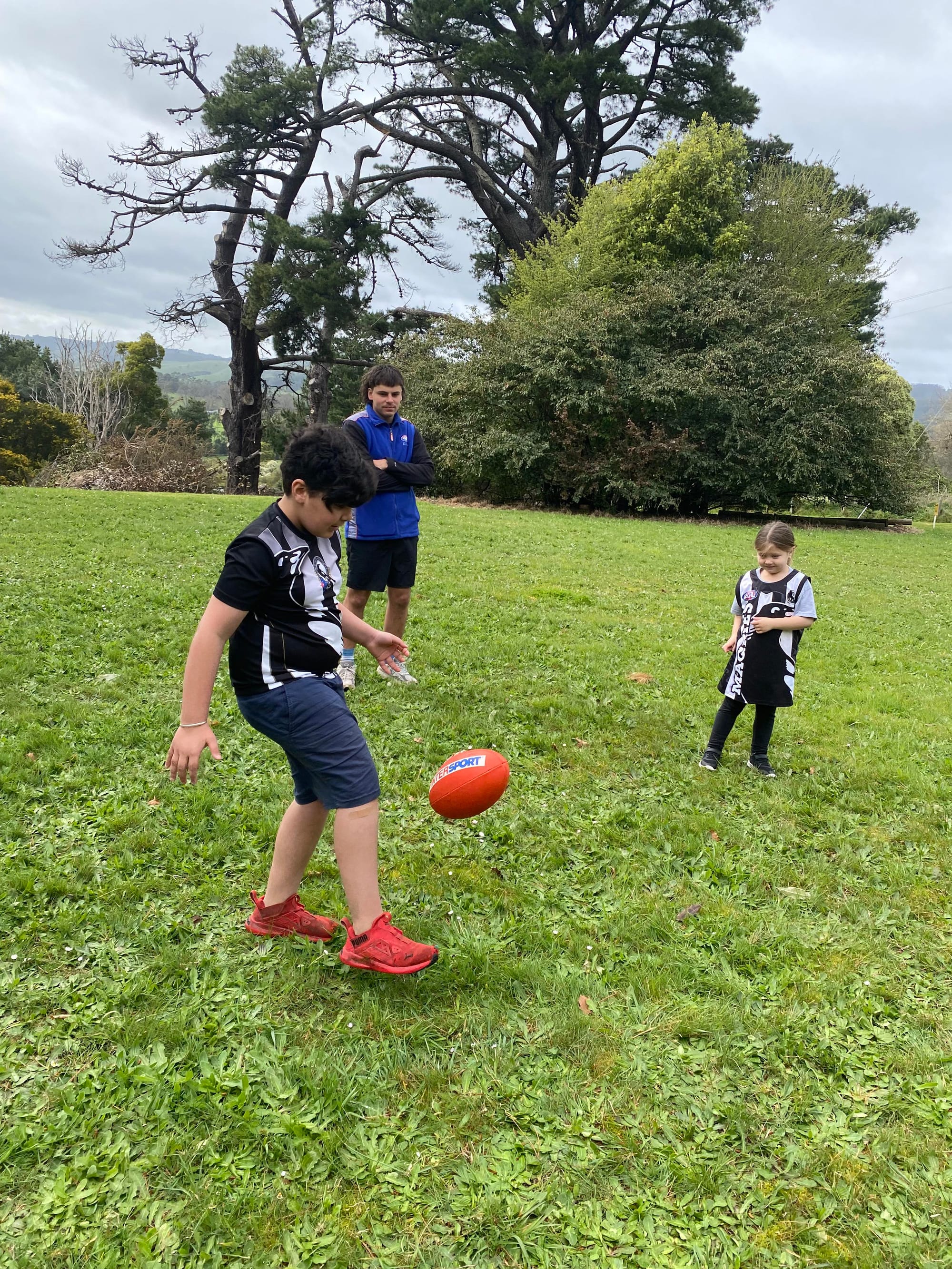 Education support Aden Guy watches as grade three Johnny Khurana and prep Madi Ashby learn to kick the footy.